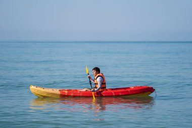 Can yeleği giymiş bir adam denizde bir kano teknesinde kürek çekiyor.