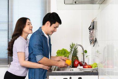 Mutlu genç çift evdeki mutfaktaki lavaboda biber ve sebze yıkıyor. Kadın sarılıyor.