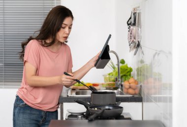 Yemek pişiren ve hazırlayan genç bir kadın evdeki mutfakta bir tablet bilgisayarın tarifine göre...