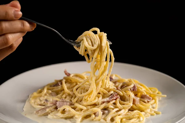 fork is spinning carbonara spaghetti on white plate on a black background