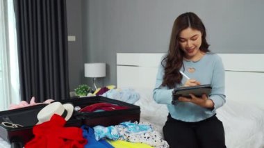 Genç bir kadın tablet yazıyor ve giysilerini bavula koyuyor evdeki bir yatakta tatil planları yapıyor..