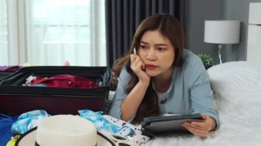 Stresli kadın tablet kullanıyor ve evdeki bir yatağa bavul yerleştiriyor. Tatil seyahati kavramı.