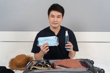 Genç adam bavul hazırlıyor. Yüz maskesi ve alkol spreyi de dahil. Bir yatakta koronavirüsü (covid-19) korumak için.