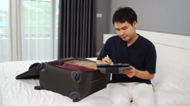 Genç adam tablet yazıyor ve giysilerini bavula koyuyor evdeki bir yatakta tatil planları yapıyor..