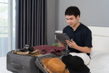 Genç adam tablet yazıyor ve giysilerini bavula koyuyor evdeki bir yatakta tatil planları yapıyor..