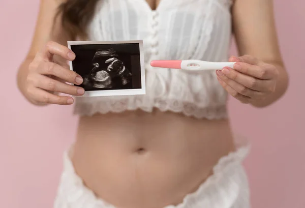 pregnant woman showing positive pregnancy test and ultrasound photo on pink background