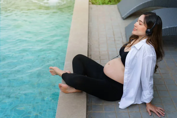 young pregnant woman sitting on edge of swimming pool while listening to music with headphone