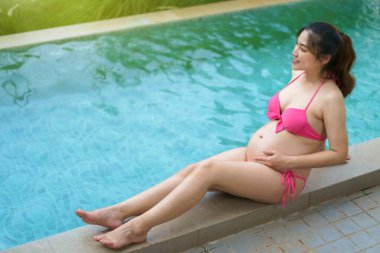 young happy pregnant woman in bikini sitting on edge of swimming pool 