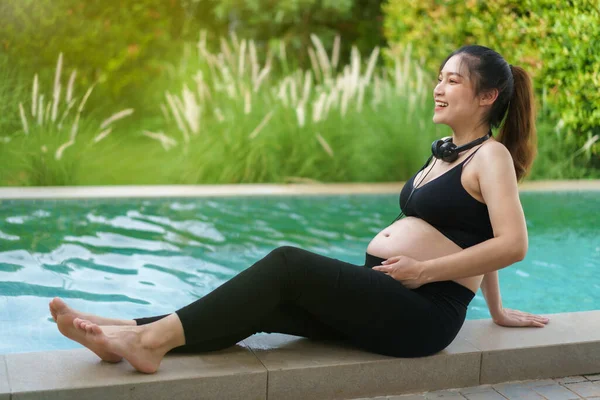 young pregnant woman sitting on edge of swimming pool while listening to music with headphone