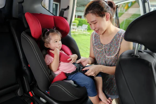 mother is fastening safety belt to unhappy toddler girl in car seat before go for ride