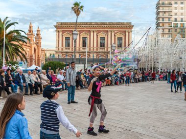 Bari, Italy - February 9, 2018: street artist making children play with big soap bubbles in a square in Bari. In the background the Margherita theatre