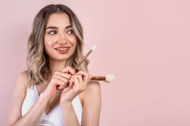 Açık pembe arka planda doğal makyajı olan güzel sarışın kız, ellerinde makyaj fırçaları, uzay kopyaları.
