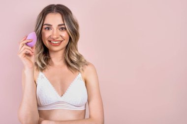 Açık pembe arka planda doğal makyajı olan güzel sarışın kız ellerinde güzellik blenderi süngeri, kopyalama alanı.