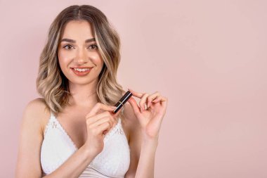 Açık pembe arka planda doğal makyajı olan güzel sarışın kız, elinde ruj, fotokopi odası var.