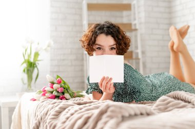 Kıvırcık saçlı esmer bir kız yatağa uzanıyor ve yüzünü bir kartla örtüyor. Yatağın üzerindeki bir buket lalenin yanında.