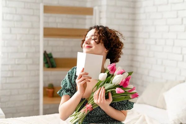 Όμορφη έκπληξη μελαχρινή κοπέλα με μια κάρτα που κρατά ένα μπουκέτο τουλίπες και κάθεται στο κρεβάτι Εικόνα Αρχείου