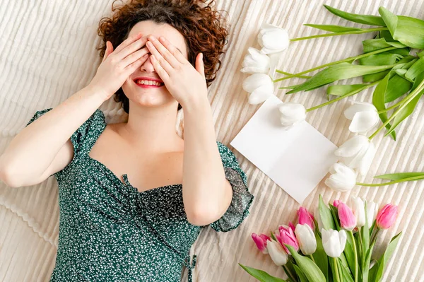 Menina feliz bonito esconde seu rosto com as mãos. deitado em uma cama com buquês de tulipas nas proximidades — Fotografia de Stock