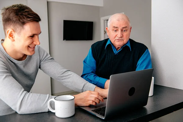 Grandson sitting on sofa with grandfather and teaching him how to use laptop. Happy family relaxing on couch at home, using modern computer together, browsing Internet, playing games, watching movies