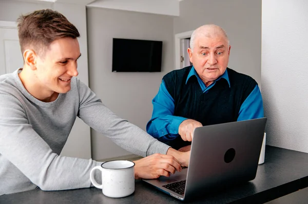 Grandson sitting on sofa with grandfather and teaching him how to use laptop. Happy family relaxing on couch at home, using modern computer together, browsing Internet, playing games, watching movies