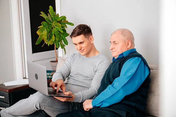 Grandson sitting on sofa with grandfather and teaching him how to use laptop. Happy family relaxing on couch at home, using modern computer together, browsing Internet, playing games, watching movies