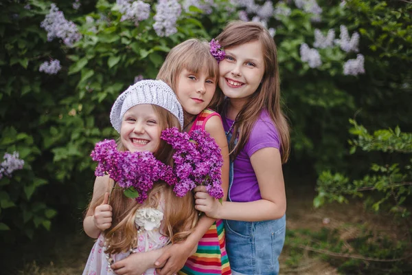 Tre sorridenti ragazze europee stanno abbracciate in un giardino estivo con fiori lilla in mano — Foto Stock