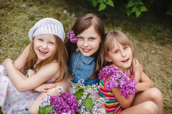 Три улыбающиеся европейские девушки сидят в саду на земле с сиреневыми цветами. Вид сверху — стоковое фото
