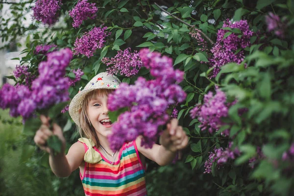 Ridendo ragazza bruna europea in un cappello beige e vestito a righe multicolore tiene i fiori lilla nelle sue mani — Foto Stock