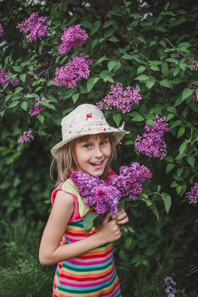 Смеющаяся брюнетка в бежевой шляпе и разноцветном полосатом платье держит в руках сиреневые цветы. — стоковое фото