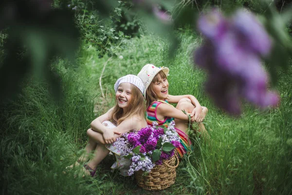 Zwei lachende europäische Mädchen mit weißen Mützen sitzen in einem Sommergarten auf dem Boden mit einem Korb bunter Flieder. — Stockfoto