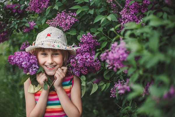 Ridendo ragazza bruna europea in un cappello beige e vestito a righe multicolore tiene i fiori lilla nelle sue mani — Foto Stock