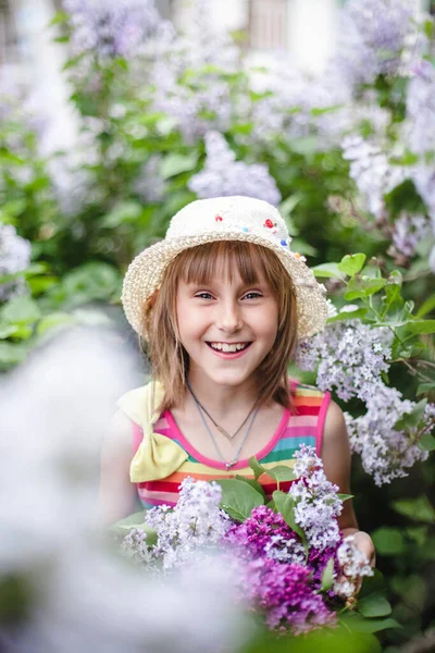 Европейская смеющаяся девушка в бежевой шляпе и полосатом платье с бантиком в сиреневом саду с корзиной сирени — стоковое фото