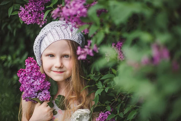 Una ragazza bionda con i capelli lunghi in un basco bianco lavorato a maglia accanto a un cespuglio di lilla tiene un ramo di lilla — Foto Stock