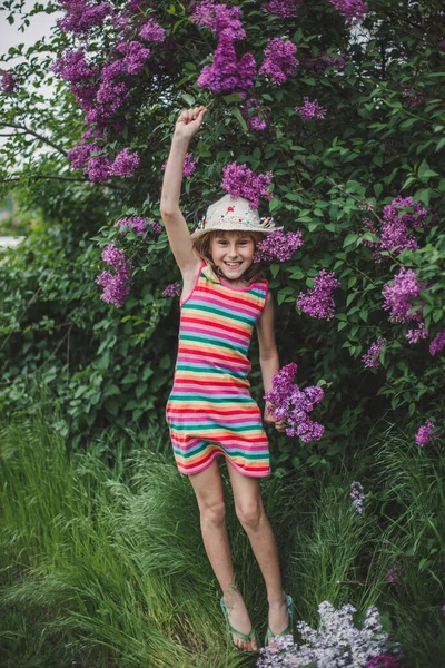 Прыгающая смеющаяся девушка европейского вида в полосатом платье и шляпе в сиреневом саду — стоковое фото