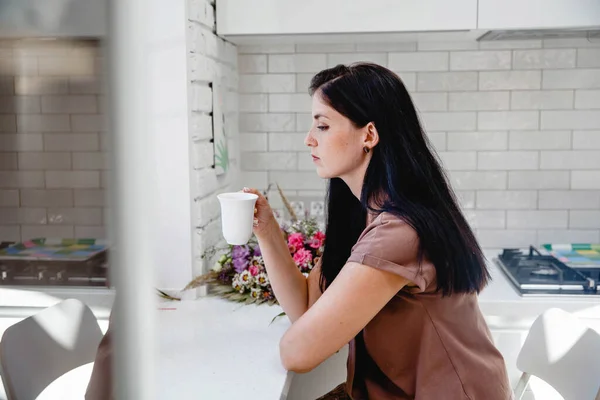 Молодая брюнетка-европейка с длинными волосами сидит за столом с цветами и пьет кофе из белой кружки — стоковое фото