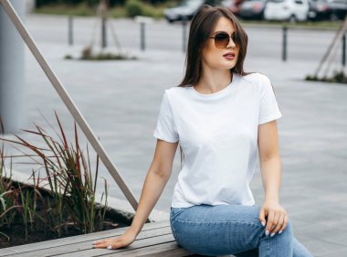 Bankta oturan şık esmer kız beyaz tişört, mavi kot pantolon ve güneş gözlüğü giyip caddeye karşı poz veriyor. Sokak fotoğrafı.
