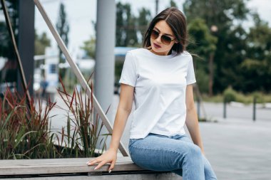 Bankta oturan şık esmer kız beyaz tişört, mavi kot pantolon ve güneş gözlüğü giyip caddeye karşı poz veriyor. Sokak fotoğrafı.