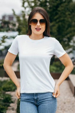 Şık esmer kız beyaz tişört, mavi kot pantolon ve güneş gözlüğü giyiyor. Sokakta poz veriyor, kentsel giyim tarzı. Sokak fotoğrafı.