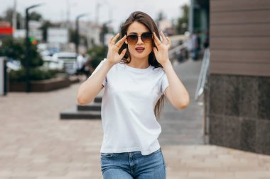 Şık esmer kız beyaz tişört, mavi kot pantolon ve güneş gözlüğü giyiyor. Sokakta poz veriyor, kentsel giyim tarzı. Sokak fotoğrafı.