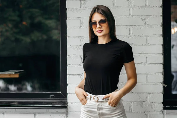 Siyah tişörtlü, güneş gözlüklü şık esmer kız sokakta poz veriyor, kentsel giyim tarzı. Sokak fotoğrafı.