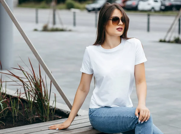 Bankta oturan şık esmer kız beyaz tişört, mavi kot pantolon ve güneş gözlüğü giyip caddeye karşı poz veriyor. Sokak fotoğrafı.