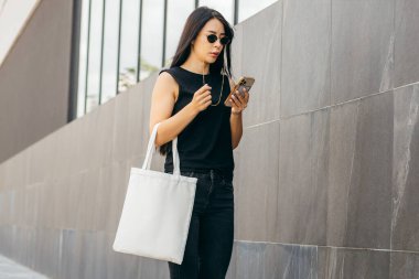 Sokakta elinde beyaz bir çanta ve cep telefonu tutan genç bir manken kız.