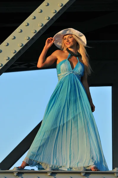 Modelo de moda en sombrero blanco y vestido de resort azul posando debajo del puente a la hora caliente del verano —  Fotos de Stock