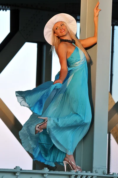 Modelo de moda em chapéu branco e vestido azul resort posando sob a ponte na hora quente de verão — Fotografia de Stock