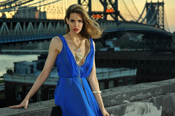 Moda modella posa sexy, indossa lungo abito da sera blu sulla posizione sul tetto con costruzione ponte in metallo su sfondo — Foto Stock