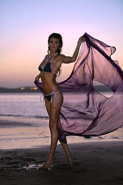Hermosa modelo de moda en bikini de diseño con tela flotante brillante de pie en la playa durante el atardecer con un fondo efectivo del cielo —  Fotos de Stock