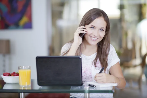 Lächelnde hübsche Frau mit Laptop, die auf dem Handy spricht und in ihr Notizbuch schreibt — Stockfoto