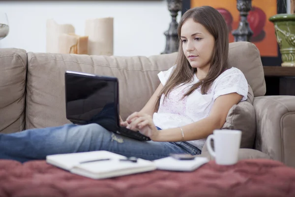 Женщина расслабилась на диване с ноутбуком, кофейной чашкой, ручкой и ноутбуком — стоковое фото