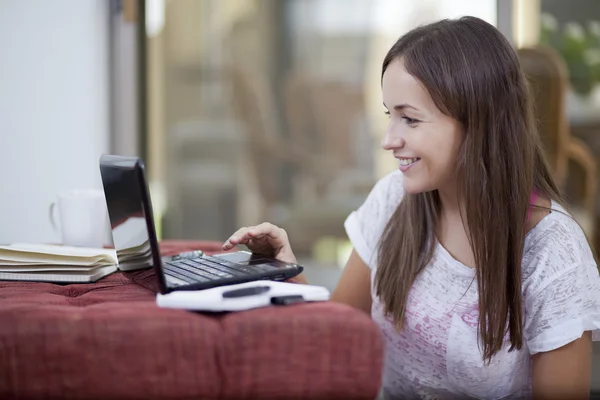 微笑着坐在地板上的红沙发旁便携式计算机上工作的年轻女子的肖像 — 图库照片