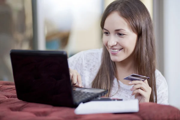 Ritratto di giovane donna sorridente che lavora su un computer portatile seduta sul pavimento accanto al divano rosso e in possesso di carta di credito — Foto Stock