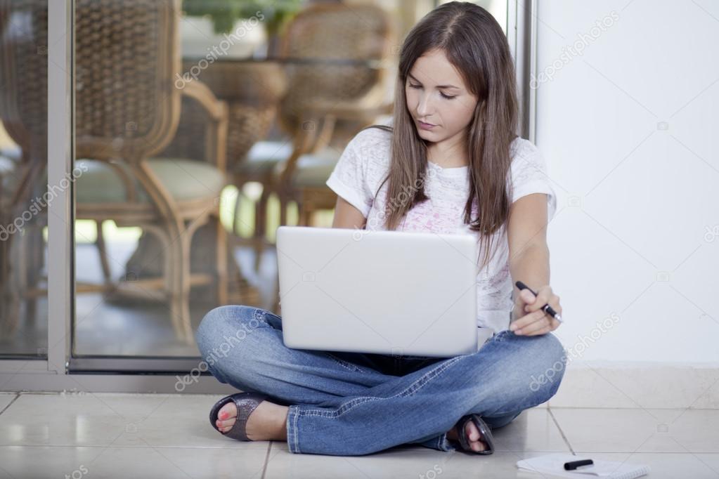 Portrait of young woman working on a laptop while sitting on the floor in front of the mirror. Cell phone, notebook and pen besides her